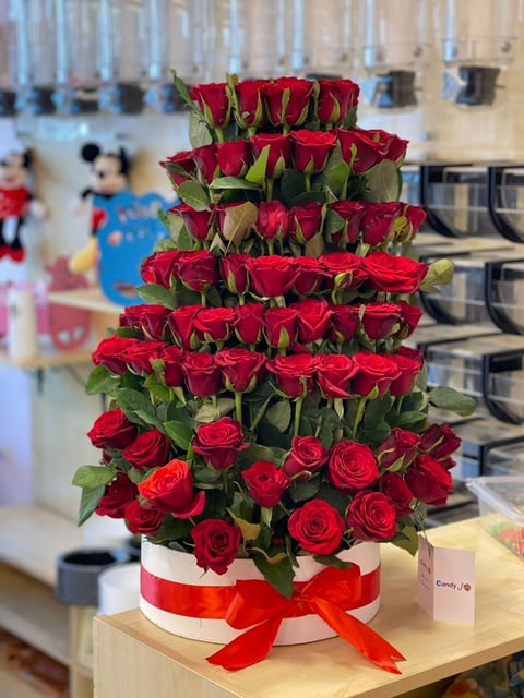 Tiered arrangement of vibrant red roses with green leaves in a white box with a red ribbon bow