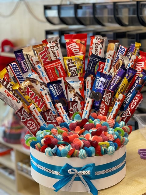 Colorful candy and chocolate bar bouquet arranged in a white box with blue ribbon, featuring various candies at the base